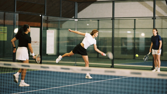 Drei Spielerinnen beim Padel auf einem Indoor-Court, eine streckt sich zum Volley